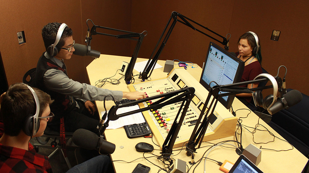 three SPOT students working in a SPOT radio production studio with microphones and an audio mixing console